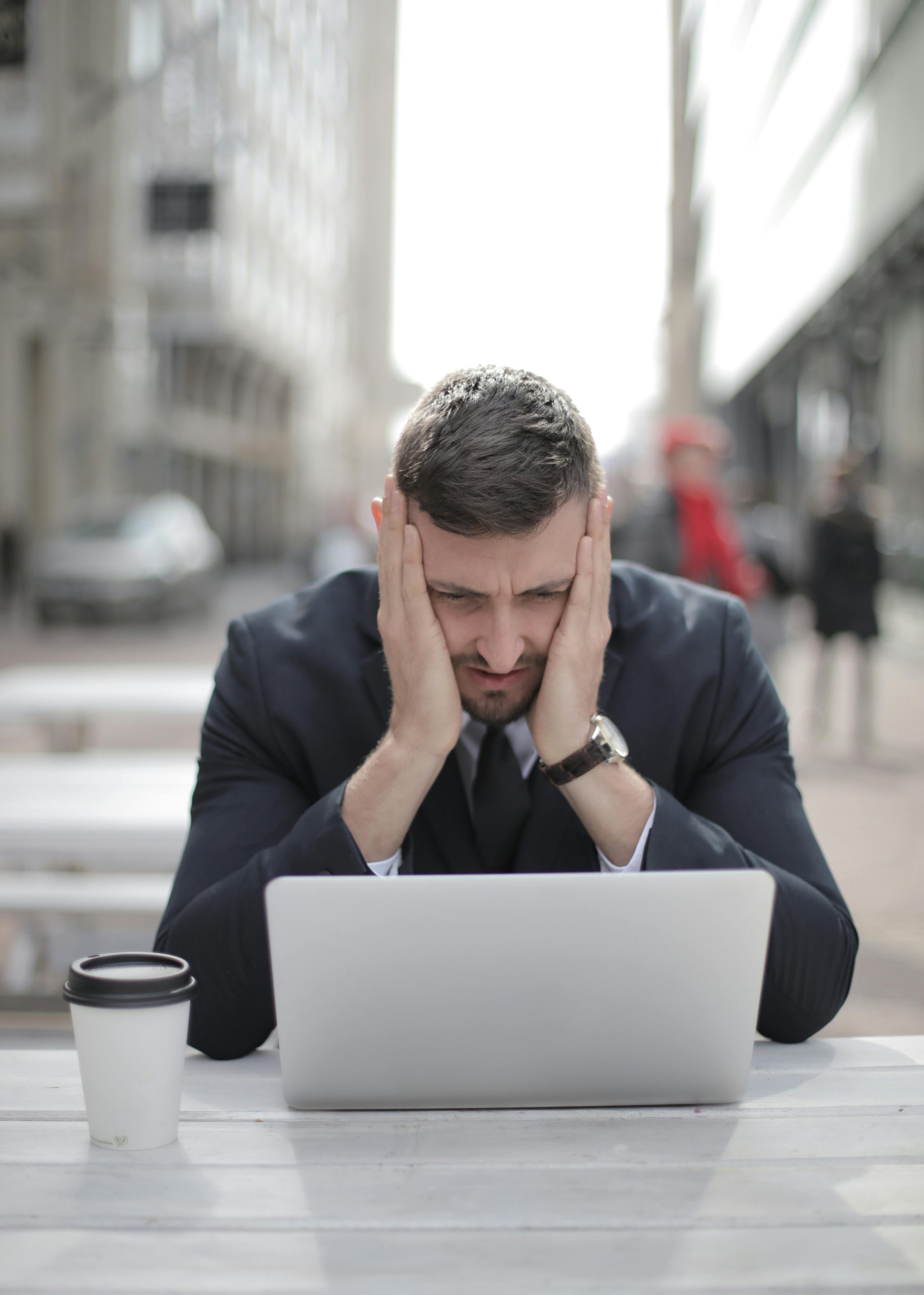 A man in a suit looks stressed while working on a laptop outdoors.